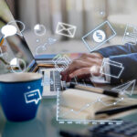 Pessoa digitando em notebook com ícones de tecnologia flutuando na tela